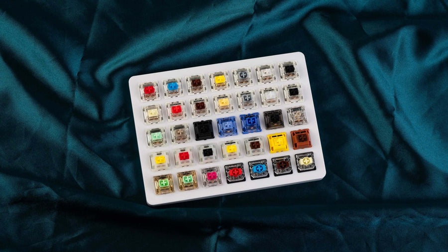 Mechanische switch tester met 35 verschillende Gateron switches in diverse kleuren en typen, gepresenteerd op een wit testboard op een satijnen achtergrond.